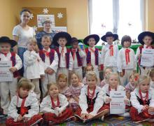 Chłopcy w czerwonych kamizelkach i kapeluszach stoją przy choince, przed nimi dziewczynki w strojach ludowych.