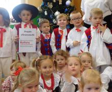 Duża grupa dzieci w tradycyjnych strojach pozuje na tle choinki i kolorowych dekoracji.